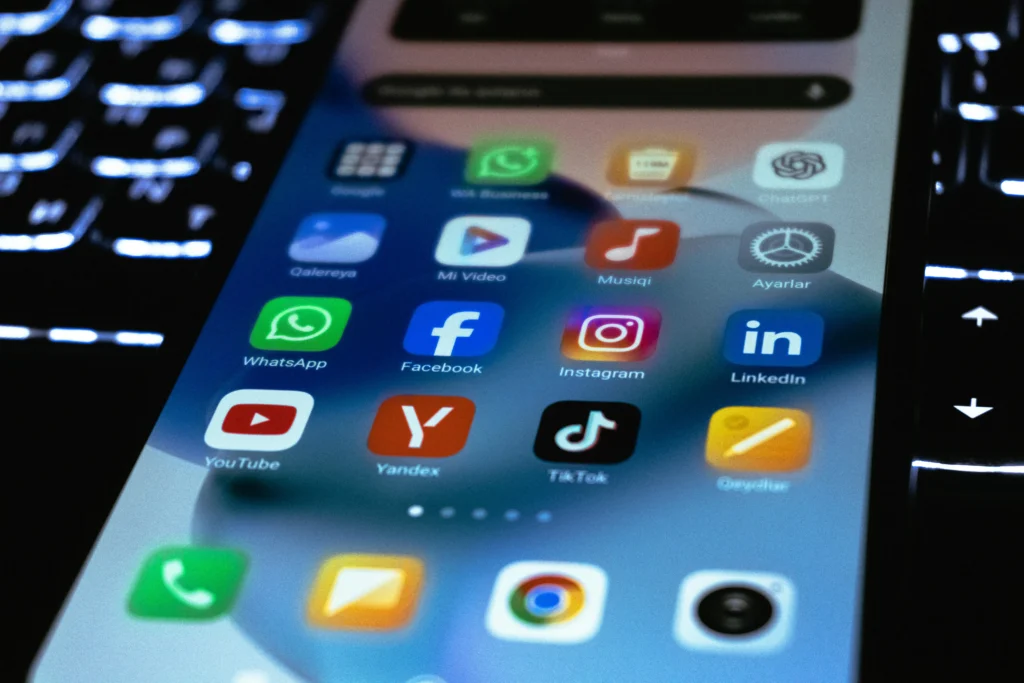 Close-up of a tilted smartphone home screen showing social and media apps: WhatsApp, Facebook, Instagram, YouTube, TikTok, LinkedIn, and more icons.