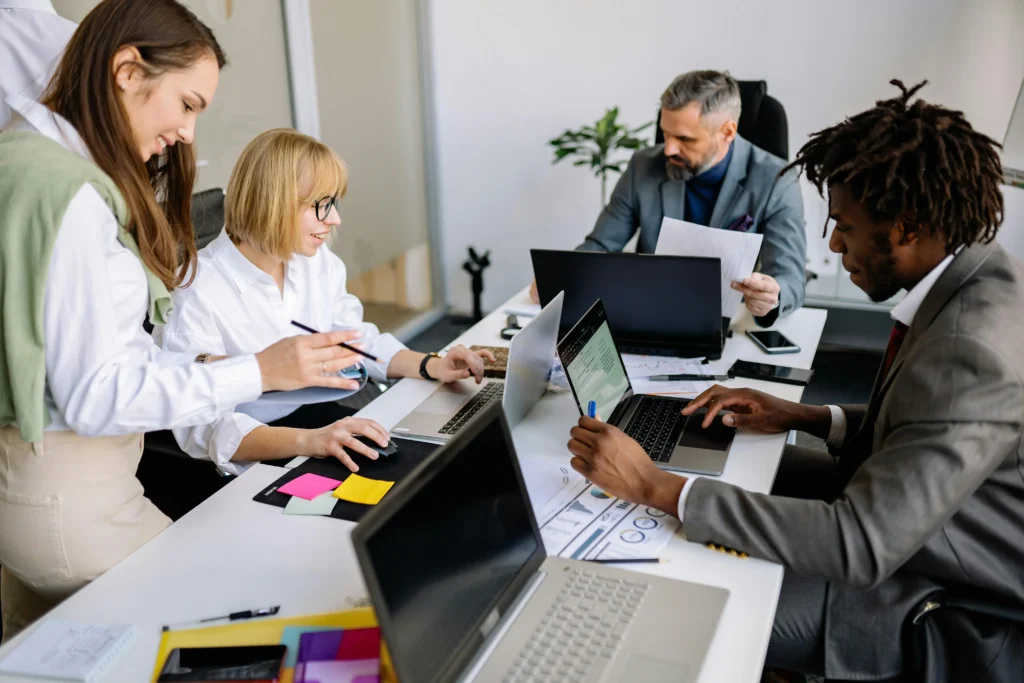 Four colleagues collaborate around a desk with laptops, papers, and colorful sticky notes in a bright office.