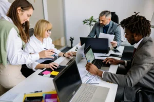 Four colleagues collaborate around a desk with laptops, papers, and colorful sticky notes in a bright office.