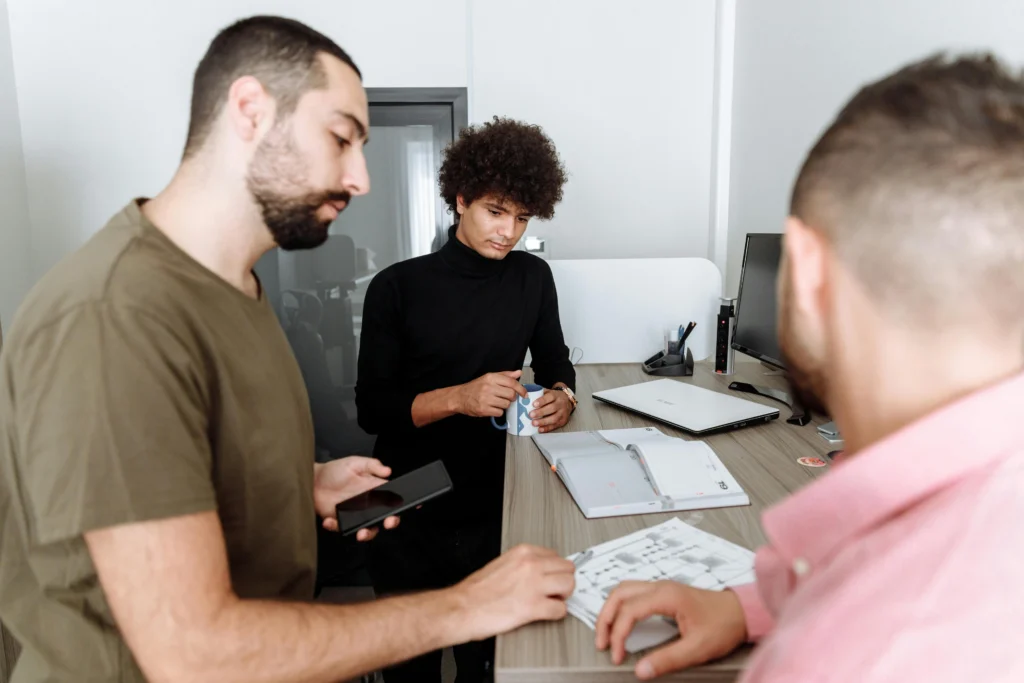 Three coworkers gather around a desk discussing documents, with one person holding a mug and another using a tablet near an open notebook and laptop.